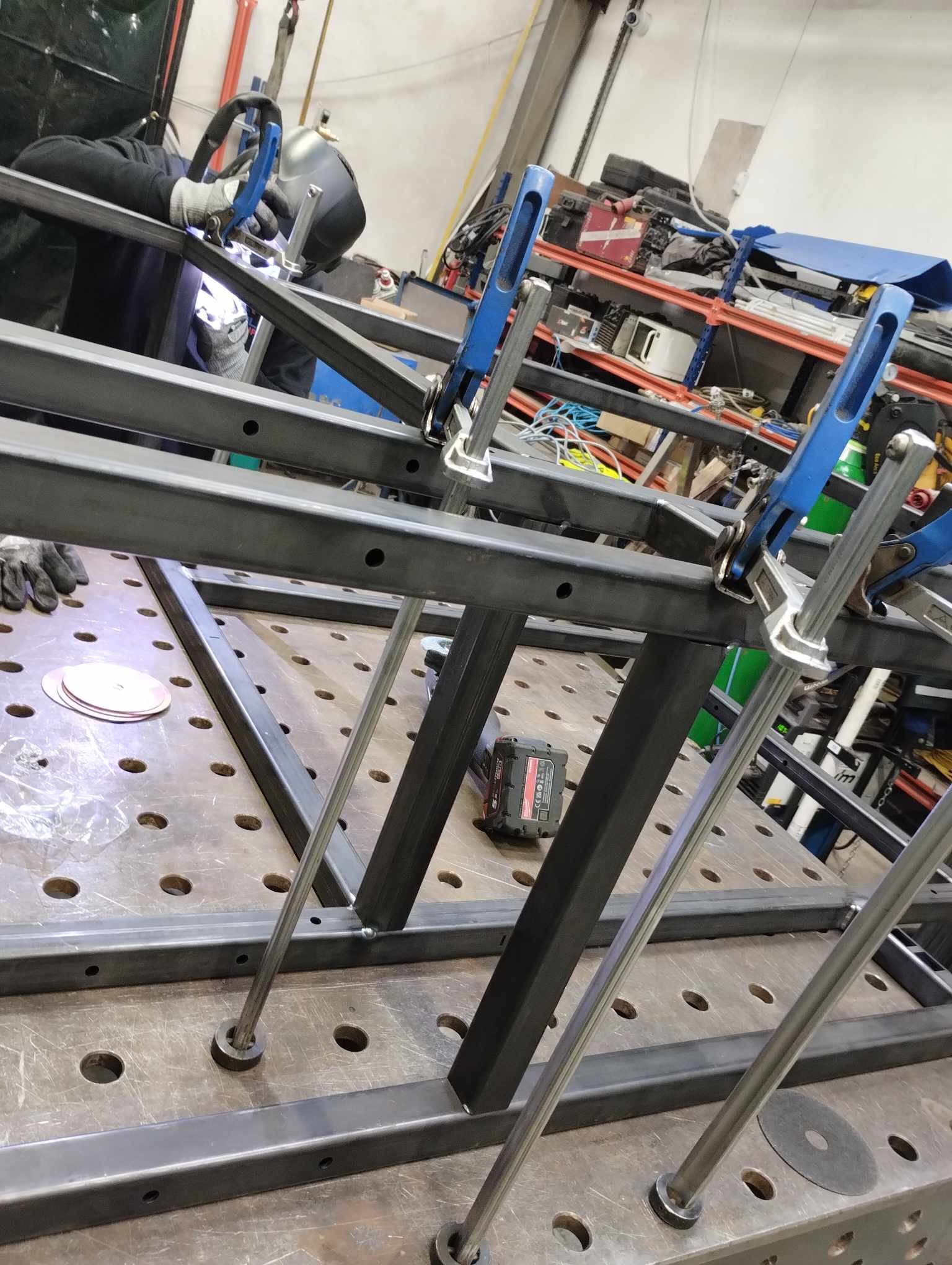 Spawanie konstrukcji stalowej z użyciem klem i maski spawalniczej, widoczny stół spawalniczy z otworami, akumulator i tarcze szlifierskie w tle warsztatu.