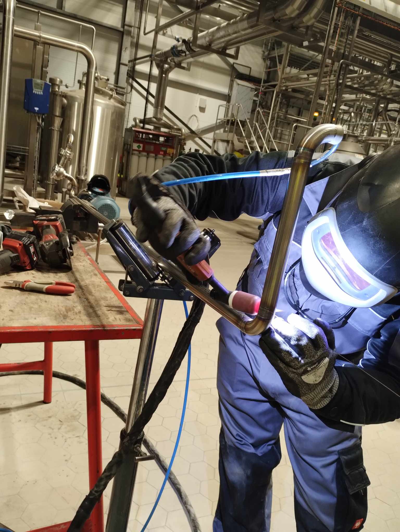 Pracownik w kombinezonie spawalniczym łączy metodą TIG rury ze stali nierdzewnej w hali przemysłowej, widoczne narzędzia i instalacje przemysłowe w tle.