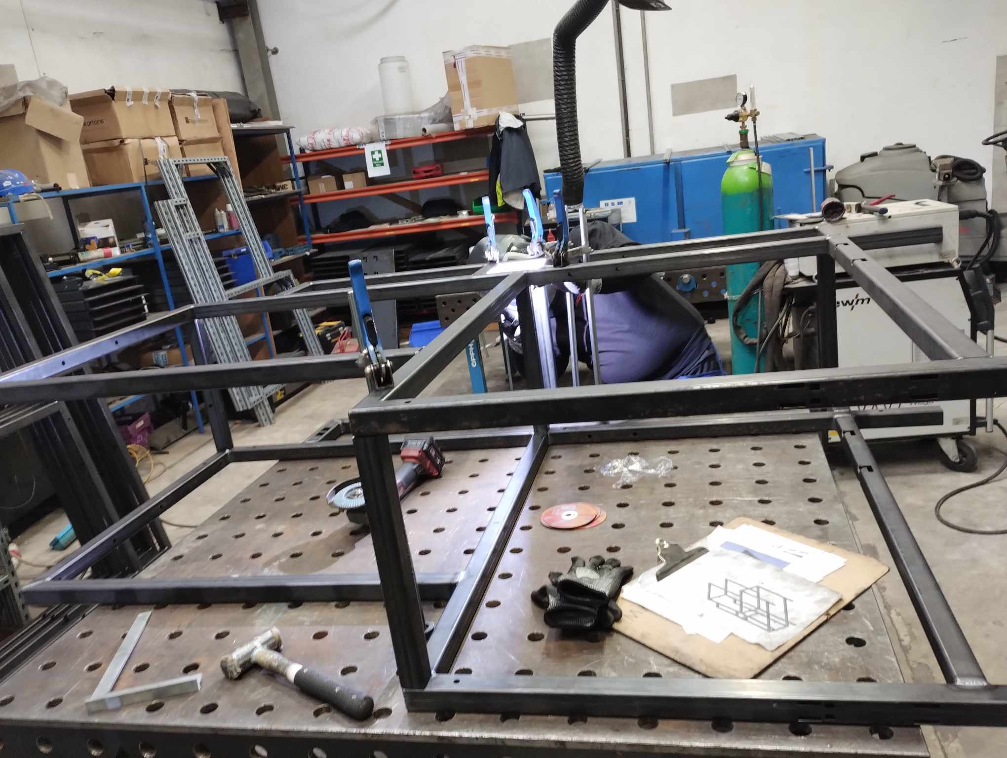 Konstrukcja stalowa w trakcie spawania na perforowanym stole spawalniczym, widoczne narzędzia, rysunek techniczny i iskry spawania