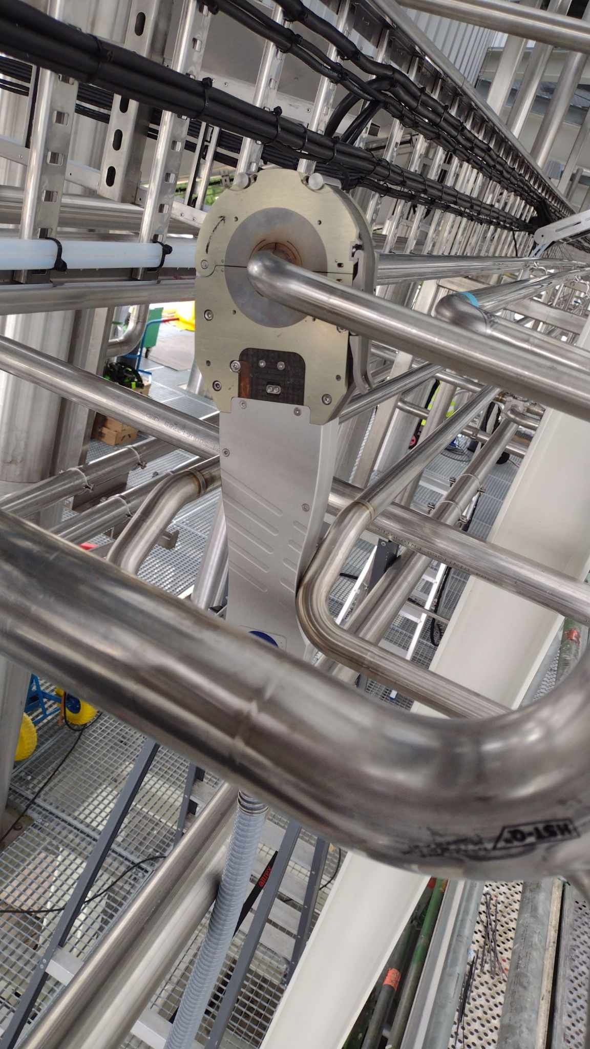 Detal metalowej konstrukcji z rur, prawdopodobnie w hali przemysłowej, widoczne elementy mocujące i przewody, perspektywa z dołu.