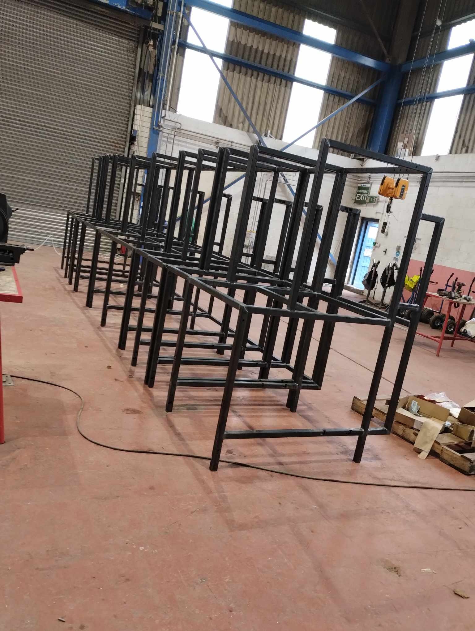 Czarne, metalowe ramy konstrukcyjne ustawione w rzędzie w hali produkcyjnej, widoczne skrzynki i narzędzia w tle.