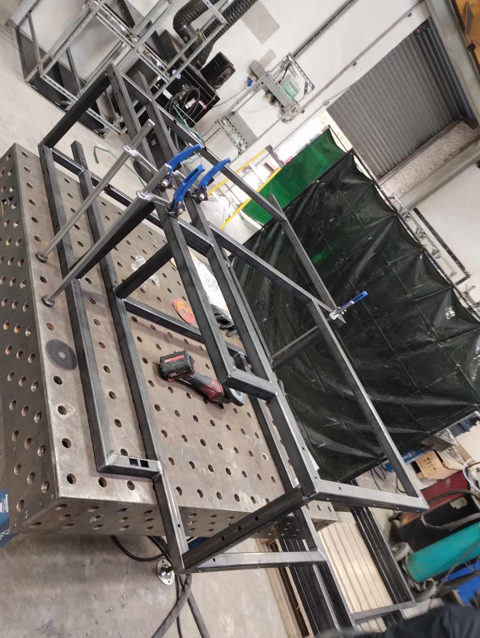 Stalowa konstrukcja spawana na perforowanym stole spawalniczym, zabezpieczona niebieskimi zaciskami, z widoczną szlifierką kątową akumulatorową i osłoną spawalniczą w tle.