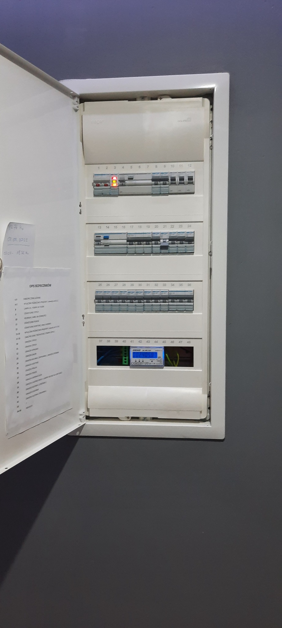 Otwarta skrzynka elektryczna z bezpiecznikami i licznikiem energii, widoczna kartka z opisem bezpieczników przytwierdzona do drzwi.
