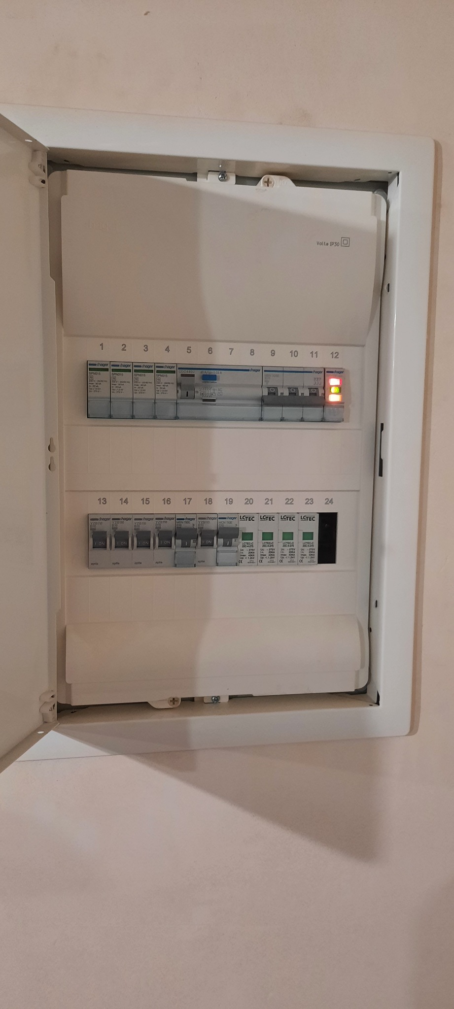 Otwarta skrzynka bezpiecznikowa, widoczne wyłączniki automatyczne różnych typów, w tym Hager i LCTEC, jeden wyłącznik w pozycji włączonej sygnalizuje diodą.