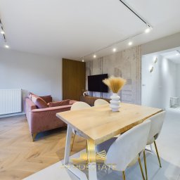 MANDU AMELIA ROGOŃ - Jasne wnętrze salonu z parkietem w jodełkę, różową sofą i drewnianym stołem z białymi krzesłami. W tle dekoracyjna ściana i telewizor. Widoczny logotyp firmy.