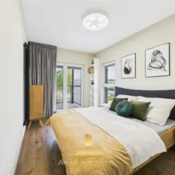 Jasna sypialnia z drewnianą podłogą i łóżkiem z szarą ramą. Balkon z widokiem na zieleń. Dwa obrazy na ścianie. Minimalistyczny wystrój.