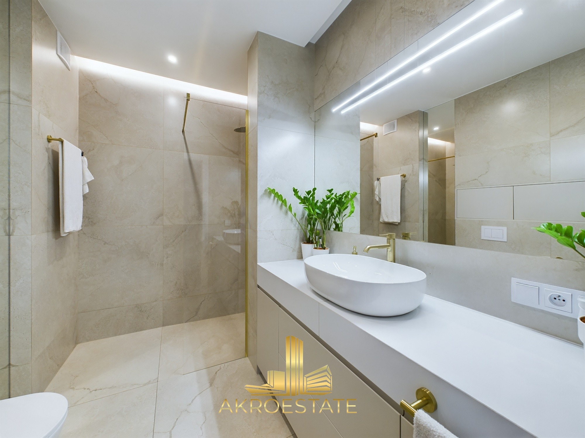 Nowoczesna łazienka z beżowymi płytkami, złotą armaturą i owalną umywalką nablatową. Widoczna kabina prysznicowa typu walk-in i roślina doniczkowa.