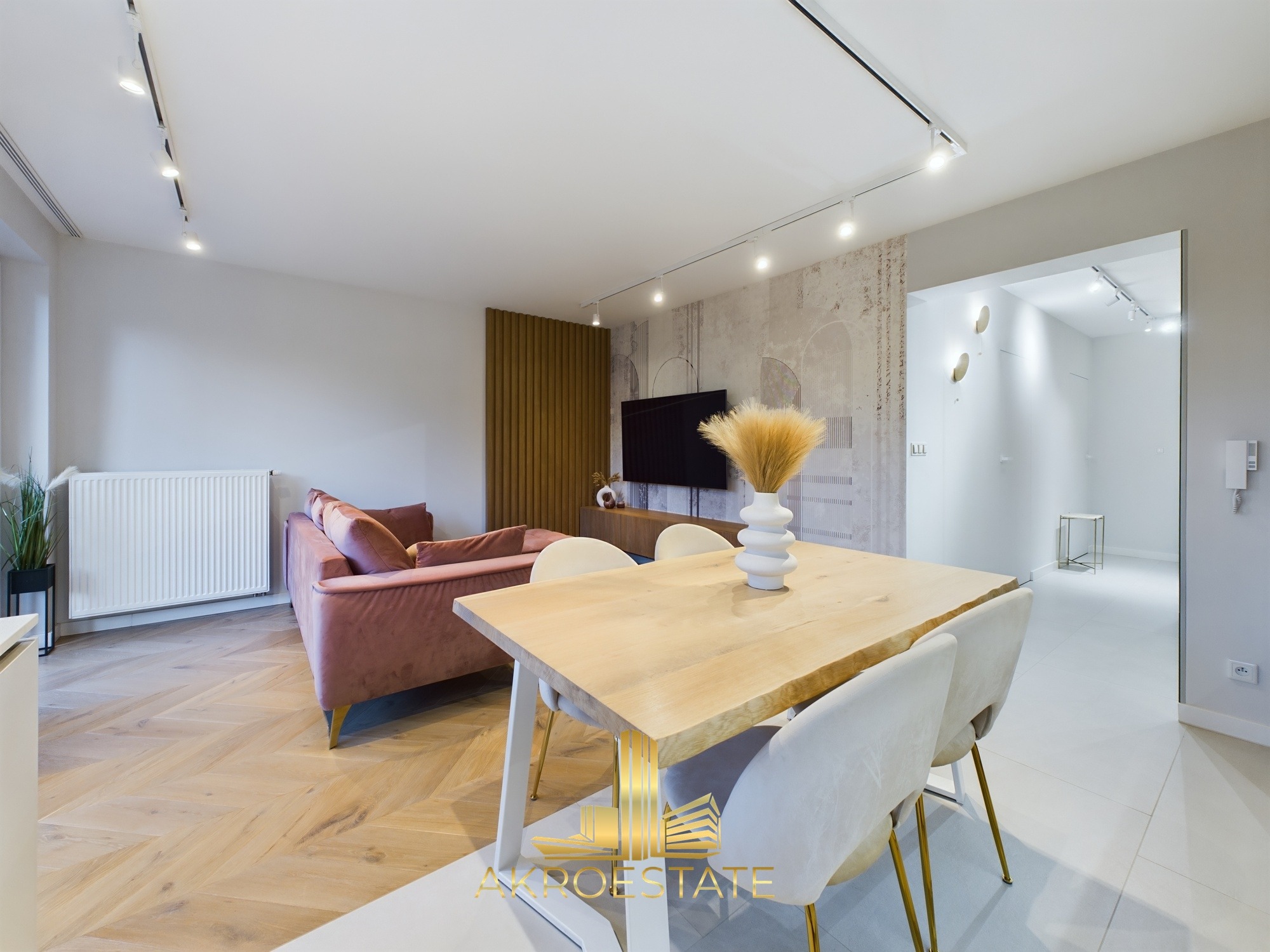 Jasne wnętrze salonu z parkietem w jodełkę, różową sofą i drewnianym stołem z białymi krzesłami. W tle dekoracyjna ściana i telewizor. Widoczny logotyp firmy.