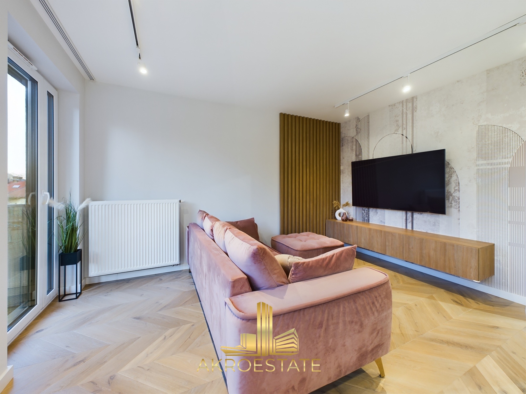 Jasny salon z parkietem w jodełkę, różową sofą i ścianą z dekoracyjnym panelem. Minimalistyczny design, nowoczesne oświetlenie punktowe i duży telewizor.