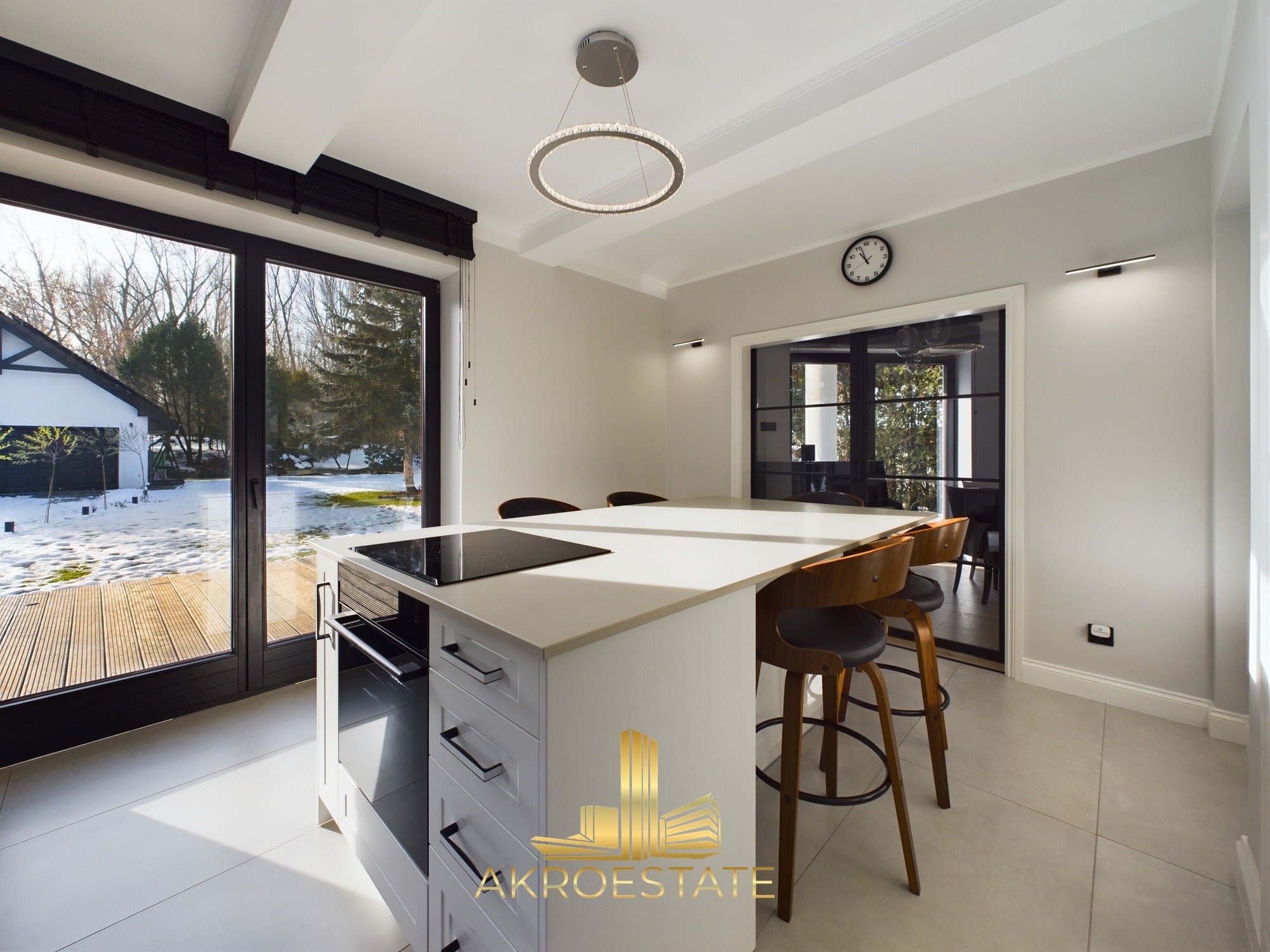 Nowoczesna kuchnia z wyspą i hokerami, widok na zaśnieżony ogród przez duże okno. Minimalistyczny design, jasne kolory, okrągła lampa sufitowa.