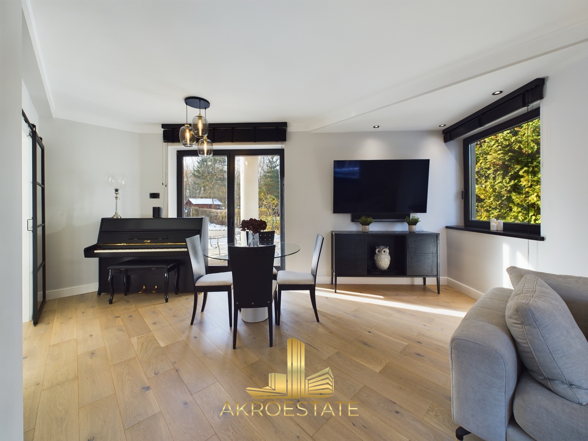 Jasne, przestronne wnętrze: parkiet, okrągły stół z krzesłami, pianino i szafka RTV z telewizorem. Za oknem zieleń. Nowoczesny design.