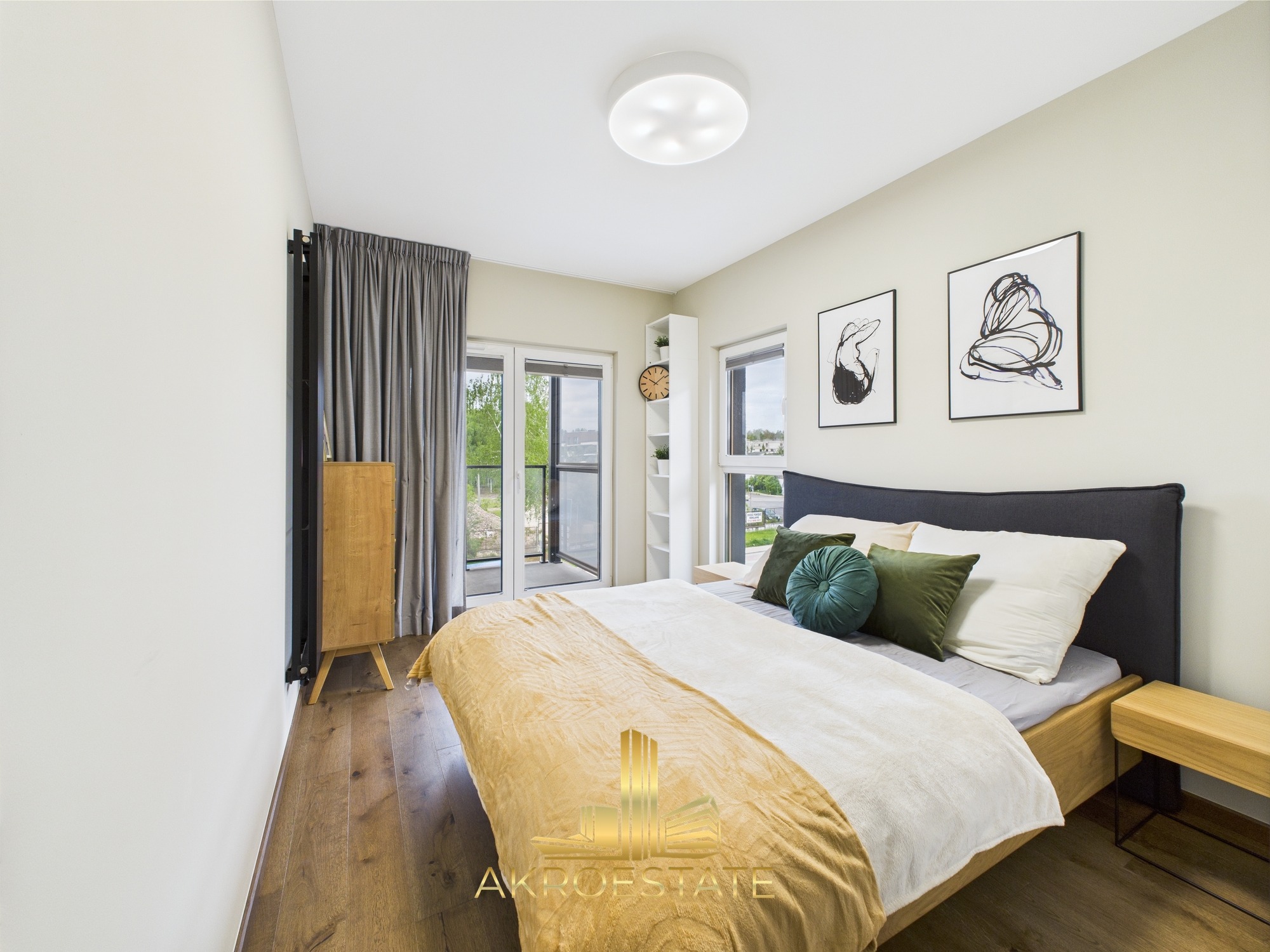 Jasna sypialnia z drewnianą podłogą i łóżkiem z szarą ramą. Balkon z widokiem na zieleń. Dwa obrazy na ścianie. Minimalistyczny wystrój.