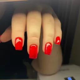 Dłoń z kwadratowymi paznokciami pomalowanymi intensywnym, błyszczącym czerwonym lakierem; widoczny fragment lampy UV do utwardzania żelu.