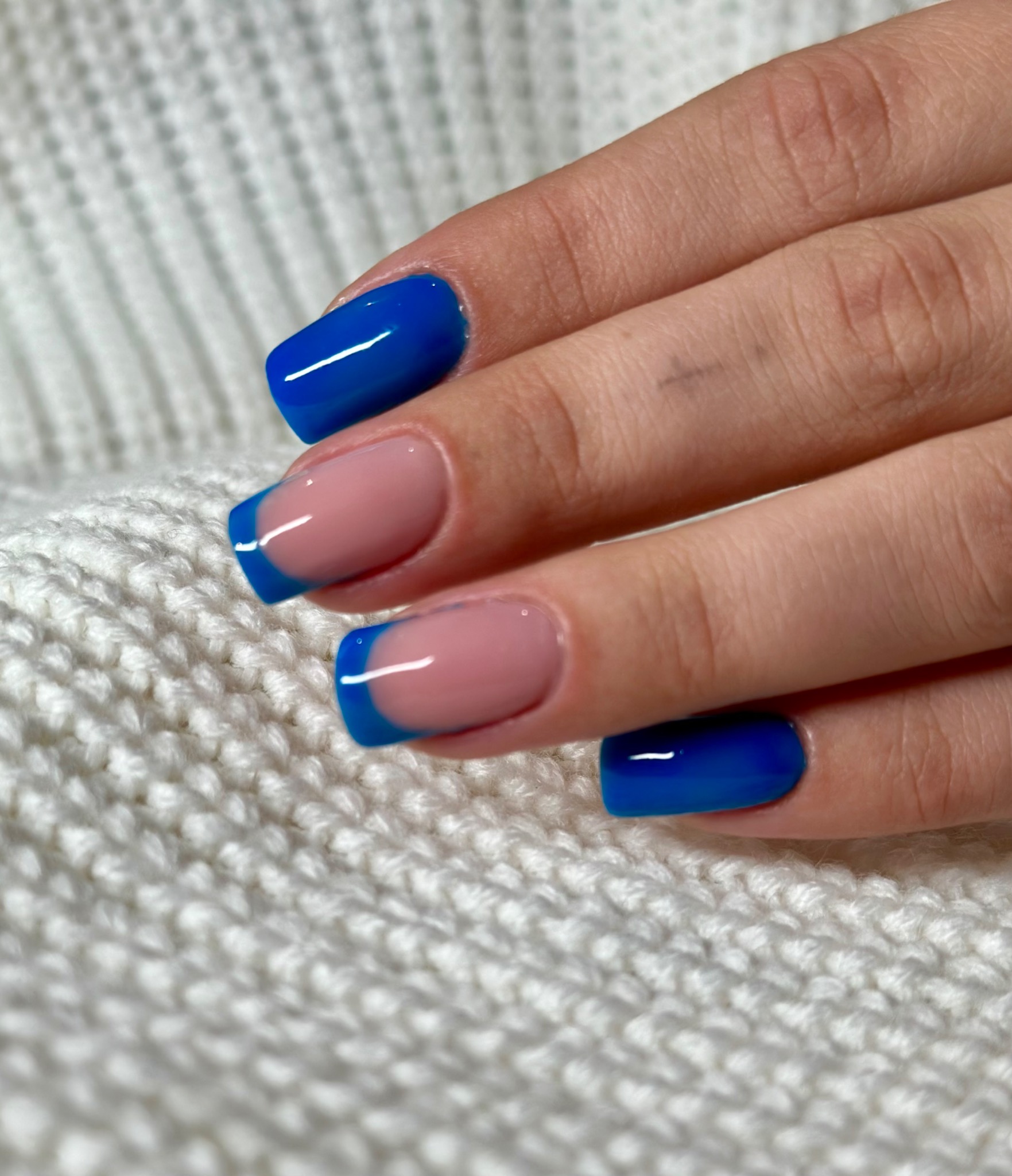 Dłoń z kwadratowymi paznokciami, jeden pomalowany na jednolity, błyszczący kobaltowy kolor, pozostałe w stylu french z kobaltowymi końcówkami, na tle białego, grubego swetra.