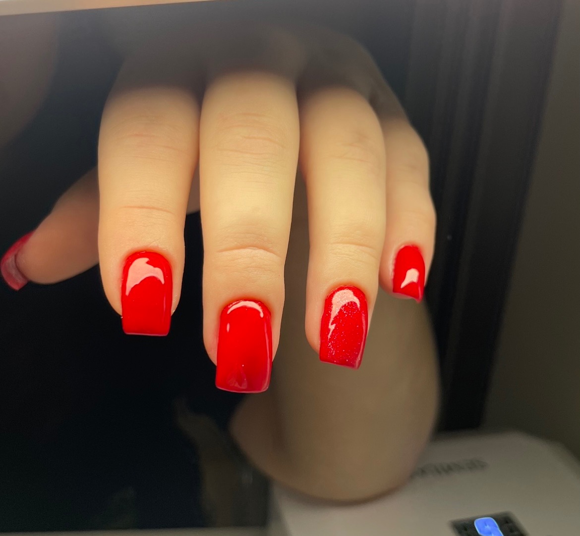 Dłoń z kwadratowymi paznokciami pomalowanymi intensywnym, błyszczącym czerwonym lakierem; widoczny fragment lampy UV do utwardzania żelu.
