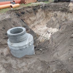Szybkop - Głęboki wykop w ziemi z ułożonymi obok betonowymi kręgami szamba. W tle widoczne rury i fragment koparki. Prace ziemne w trakcie realizacji.