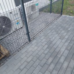 Szybkop - Ułożona szara kostka brukowa, obok ogrodzenie panelowe i klimatyzatory na żwirowym podłożu. Widok z góry, ukośny kadr.