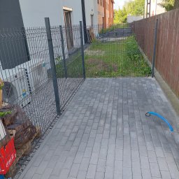 Szybkop - Ułożona szara kostka brukowa na podwórku z widocznym ogrodzeniem panelowym i fragmentem niezagospodarowanego terenu z boku. W tle budynki w trakcie budowy.