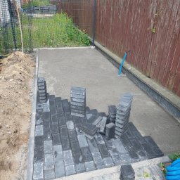 Szybkop - Częściowo ułożona, grafitowa kostka brukowa na podsypce. Stosy kostki obok ułożonego fragmentu. W tle drewniany płot i trawa. Widoczny niebieski przewód.