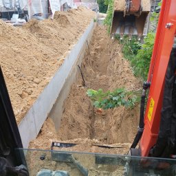 Szybkop - Wąski wykop z betonową ścianą, widok z kabiny koparki. Na dnie wykopu widoczne rury i gałąź. Koparka w trakcie pracy, ziemia usypana po bokach wykopu.