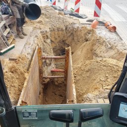 Szybkop - Głęboki wykop zabezpieczony szalunkiem stalowym. Robotnik w kasku i rękawicach umieszcza rurę w wykopie. Widok z kabiny koparki, w tle ulica z przejściem dla pieszych.