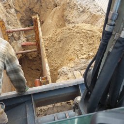 Szybkop - Głęboki wykop zabezpieczony szalunkiem stalowym, widoczne rury kanalizacyjne, fragment koparki i pracownik w koszuli w kratę. Prace ziemne w toku.