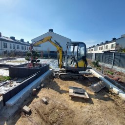 Szybkop - Żółta mini koparka na placu budowy ogrodu, w tle zabudowa szeregowa. Widoczne obrzeża rabat, kostka brukowa i przygotowane miejsce pod nasadzenia.