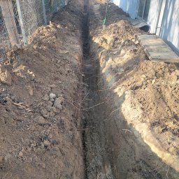 Szybkop - Wąski wykop w ziemi, widoczne korzenie, po prawej stronie budynku. Na górze kadru siatka ogrodzeniowa i zielona łopata wbita w ziemię. Słoneczny dzień.