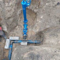 Szybkop - Niebieska instalacja wodociągowa w wykopie ziemnym, zabezpieczona betonowymi bloczkami. Widoczne złącza i rury. Fragment buta i spodni osoby w lewym dolnym rogu.