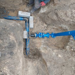 Szybkop - Niebieskie rury wodociągowe w wykopie, podparte betonowymi bloczkami. Widoczne złącza i zawory. Osoba w roboczym ubraniu w tle. Prace instalacyjne w ziemi.
