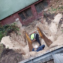 Szybkop - Głęboki wykop zabezpieczony metalowymi szalunkami, dwóch robotników w środku, jeden w kamizelce odblaskowej, drugi w kasku, prace ziemne przy budynku.