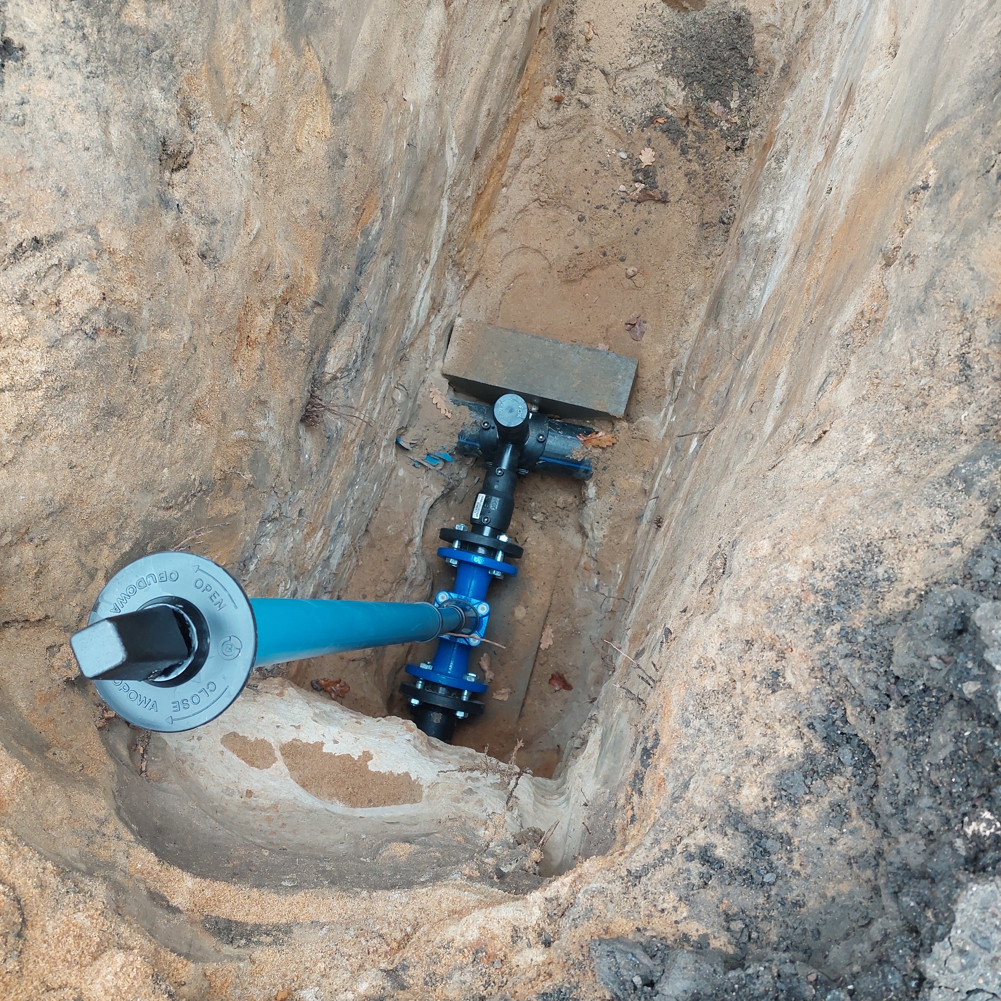 Niebieska rura wodociągowa z zaworem w wykopie z piasku, widok z góry. Elementy instalacji połączone śrubami. Na górze wykopu leży szara cegła.