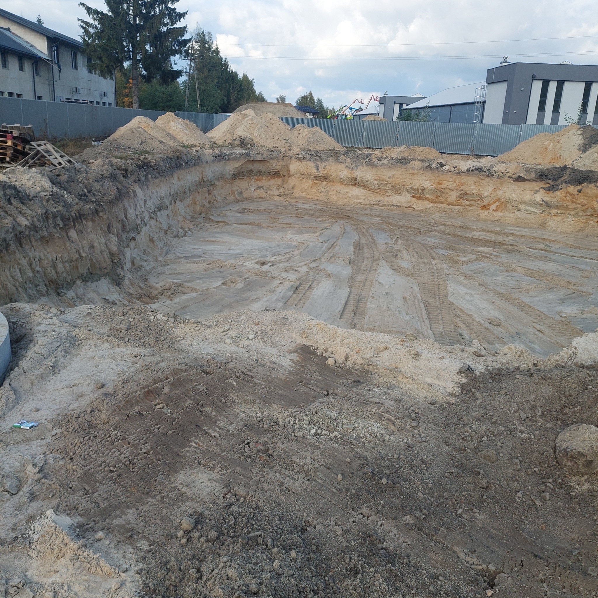 Szeroki wykop pod fundamenty z widocznymi śladami opon maszyn budowlanych na dnie, otoczony usypaną ziemią i budynkami w tle.
