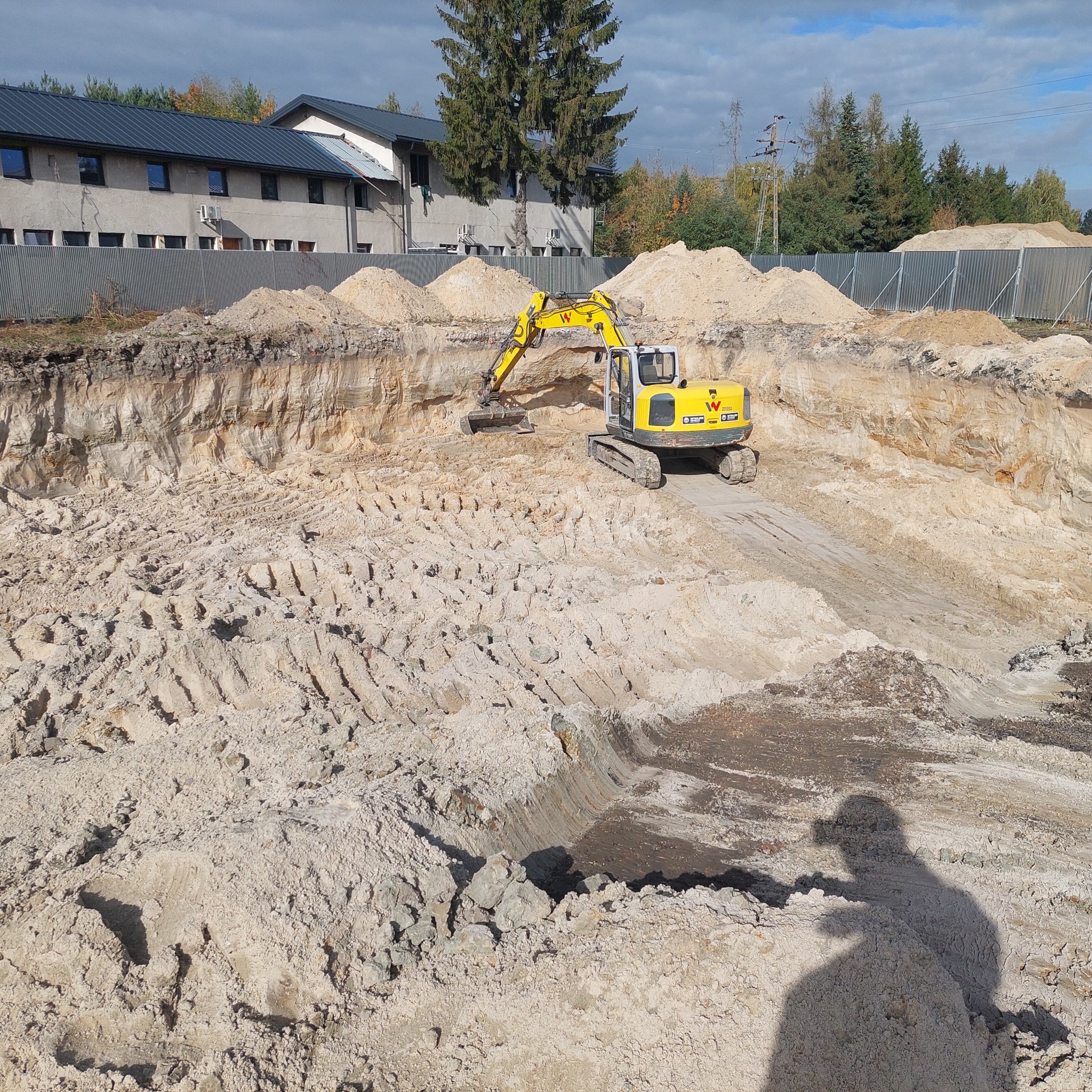 Żółta koparka Wacker Neuson w wykopie, przygotowuje teren pod budowę. Widoczne warstwy ziemi, hałdy piasku i cień osoby robiącej zdjęcie. Praca w toku.