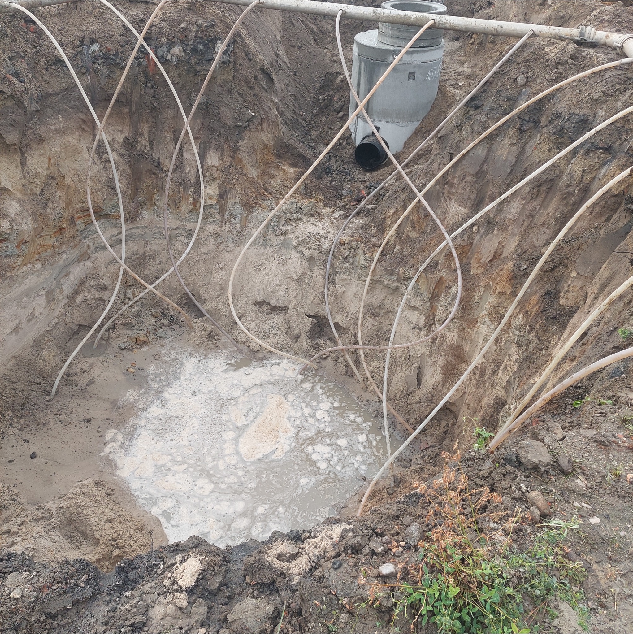 Głęboki wykop z betonowym zbiornikiem i siecią rur, częściowo wypełniony wodą. Widoczne ślady prac ziemnych i instalacyjnych.