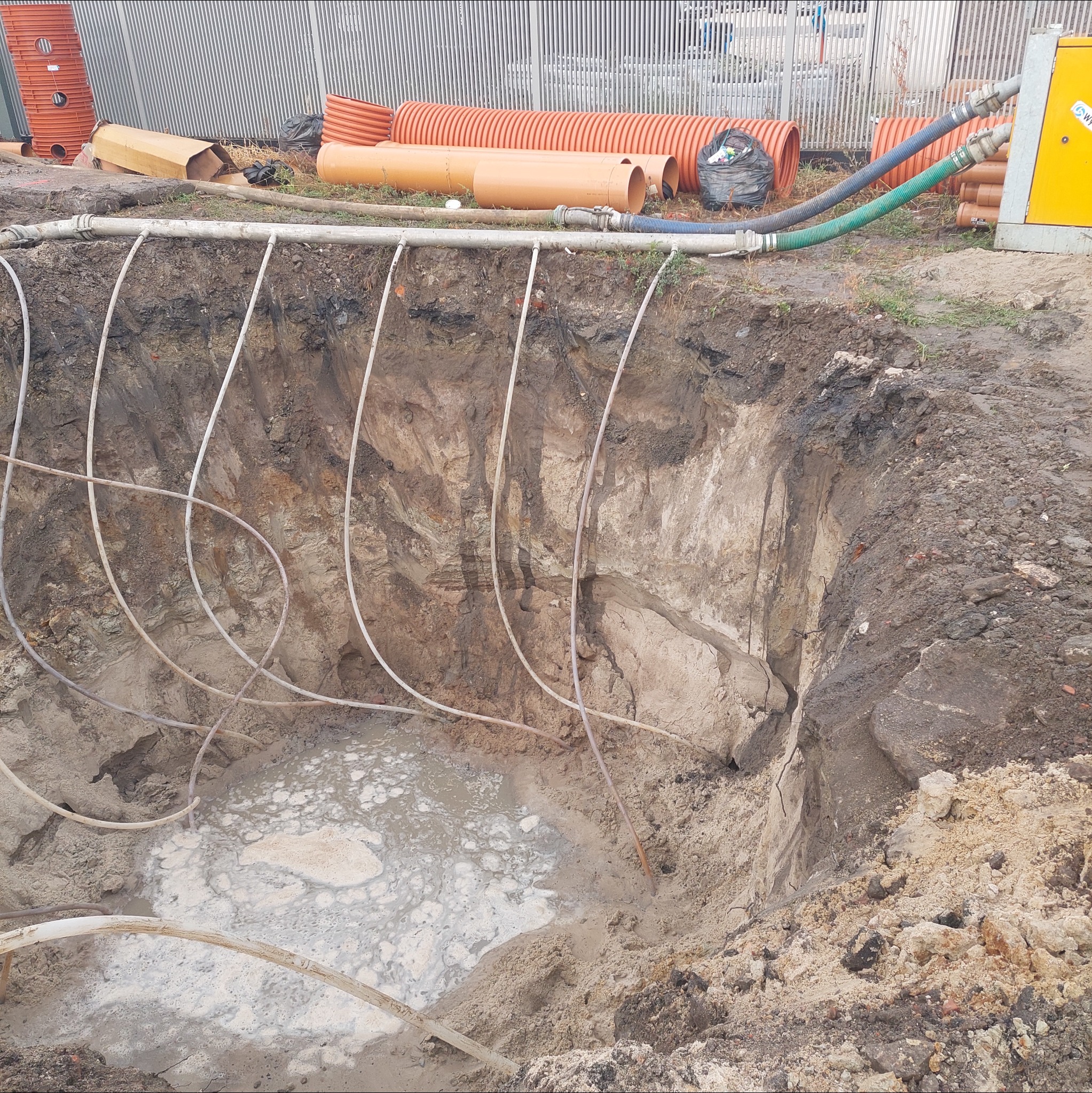 Głęboki wykop z instalacją odwadniającą, pompy i rury na powierzchni. Widoczne ślady wody w dole wykopu. Szare tło z metalowym ogrodzeniem.