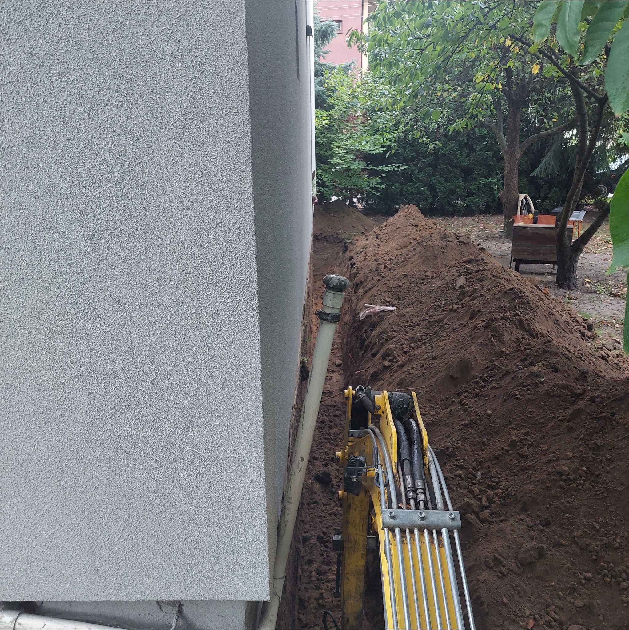 Wąski wykop wzdłuż ściany budynku, widoczna rura i fragment żółtej koparki. Ziemia usypana wzdłuż wykopu, w tle zieleń drzew.