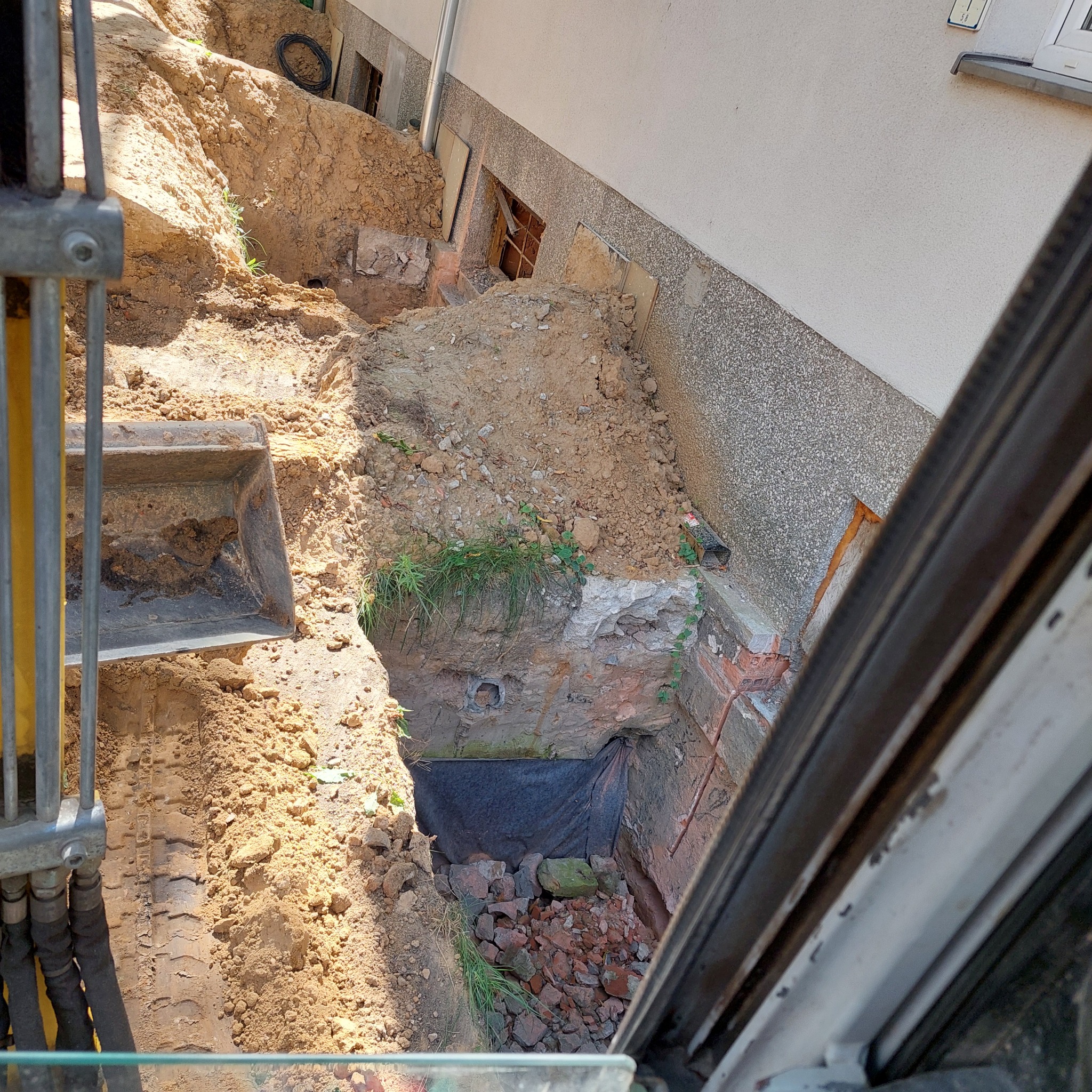 Głęboki wykop ziemny przy ścianie budynku, widok z kabiny koparki. W dole gruz i czarna folia. Ściany wykopu z ziemi i cegieł. Widoczne okna piwniczne.