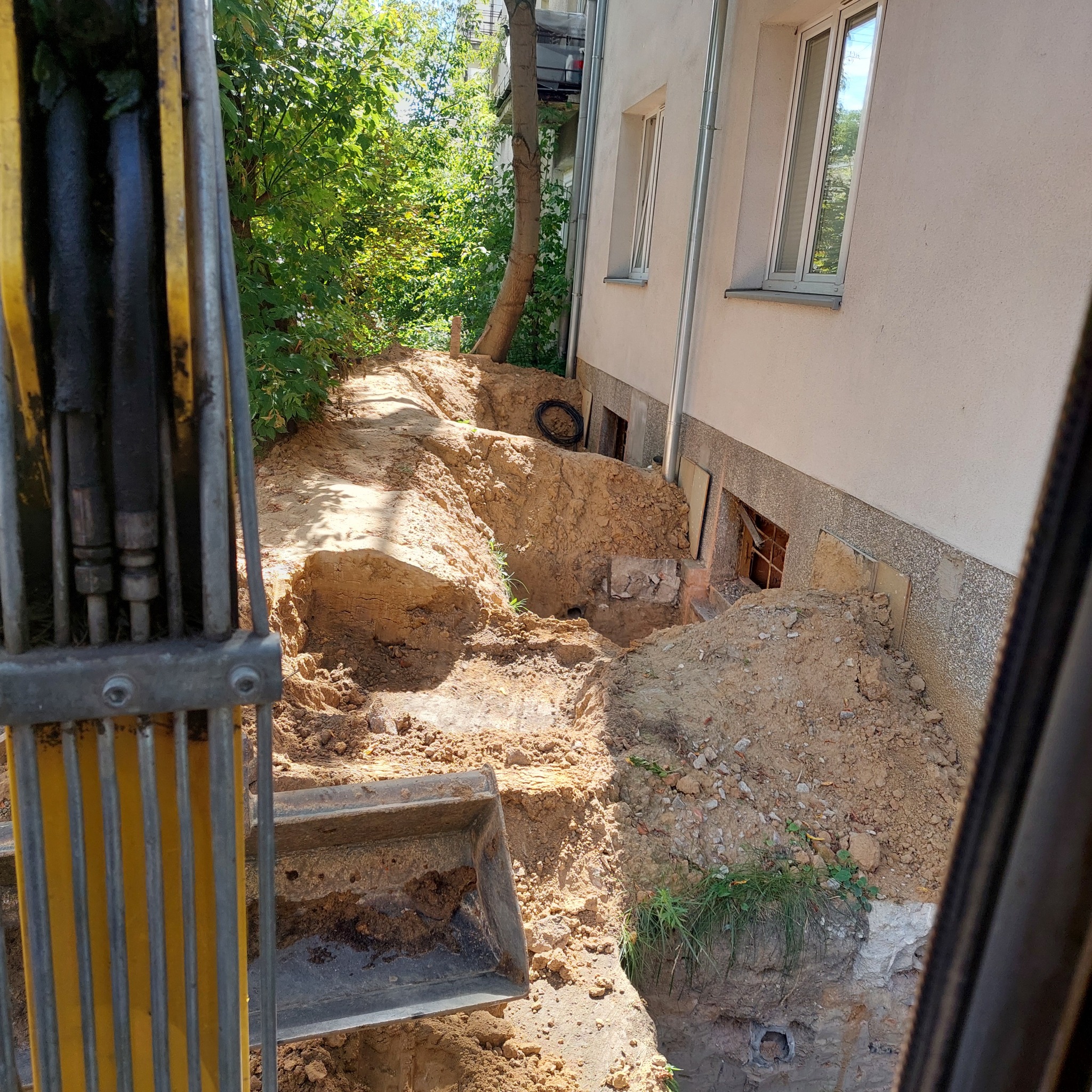 Wykop ziemny przy budynku mieszkalnym, widoczny z kabiny koparki. Wykop odsłania fundamenty i piwniczne okienka. Obok rośnie drzewo.