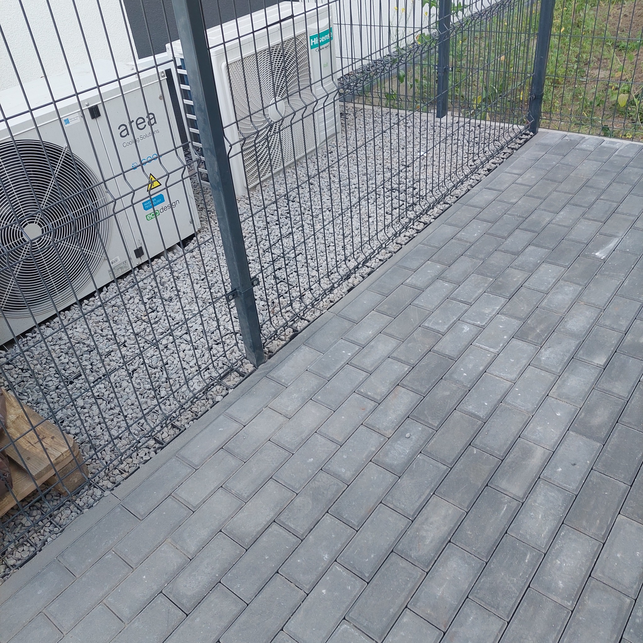 Ułożona szara kostka brukowa, obok ogrodzenie panelowe i klimatyzatory na żwirowym podłożu. Widok z góry, ukośny kadr.