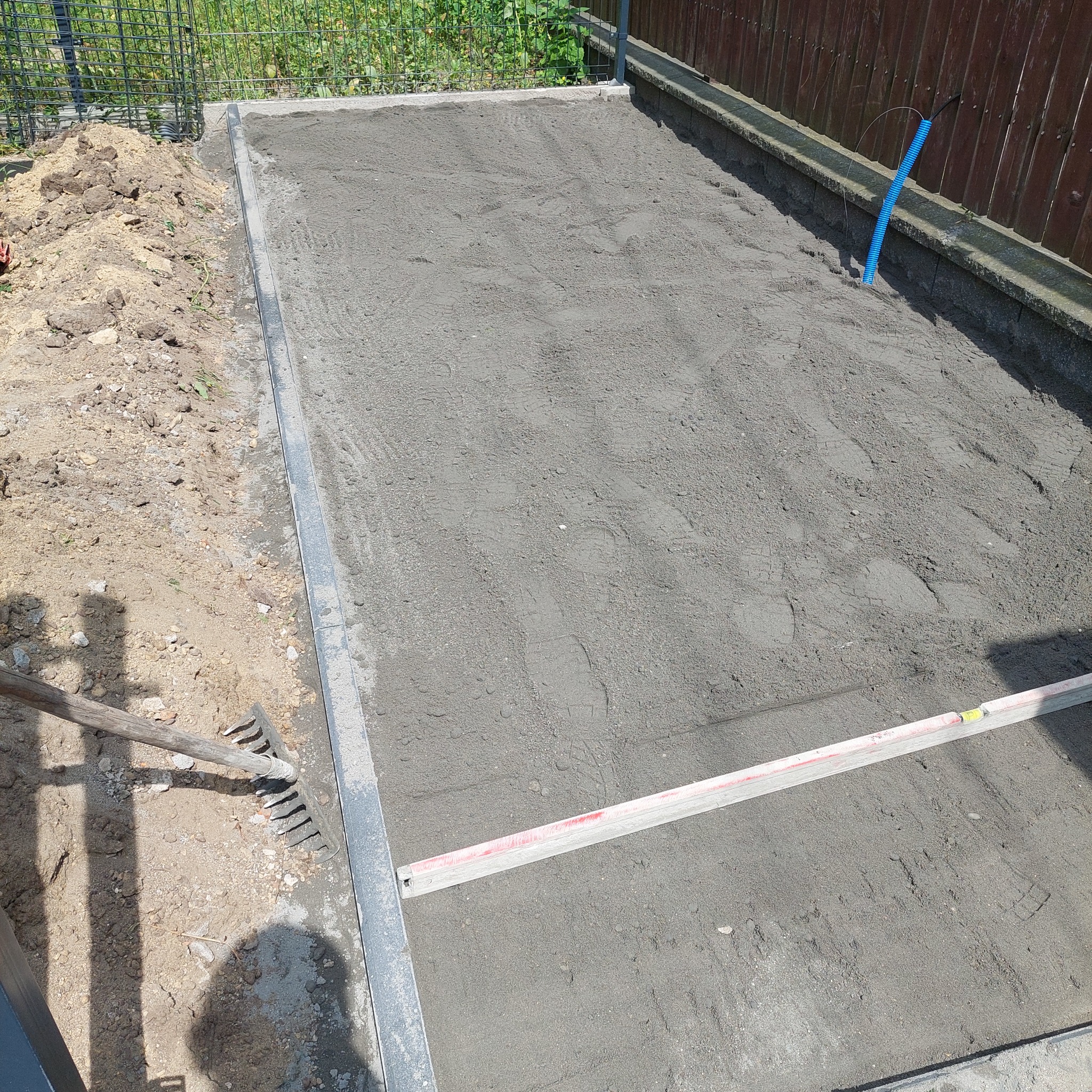 Przygotowany teren pod kostkę brukową z widocznym poziomowaniem, obrzeżami i narzędziami. W tle ogrodzenie z siatki i drewniany płot.