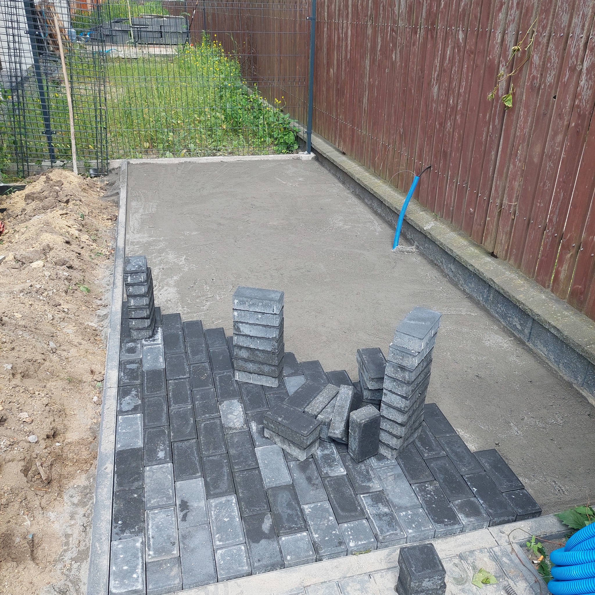 Częściowo ułożona, grafitowa kostka brukowa na podsypce. Stosy kostki obok ułożonego fragmentu. W tle drewniany płot i trawa. Widoczny niebieski przewód.