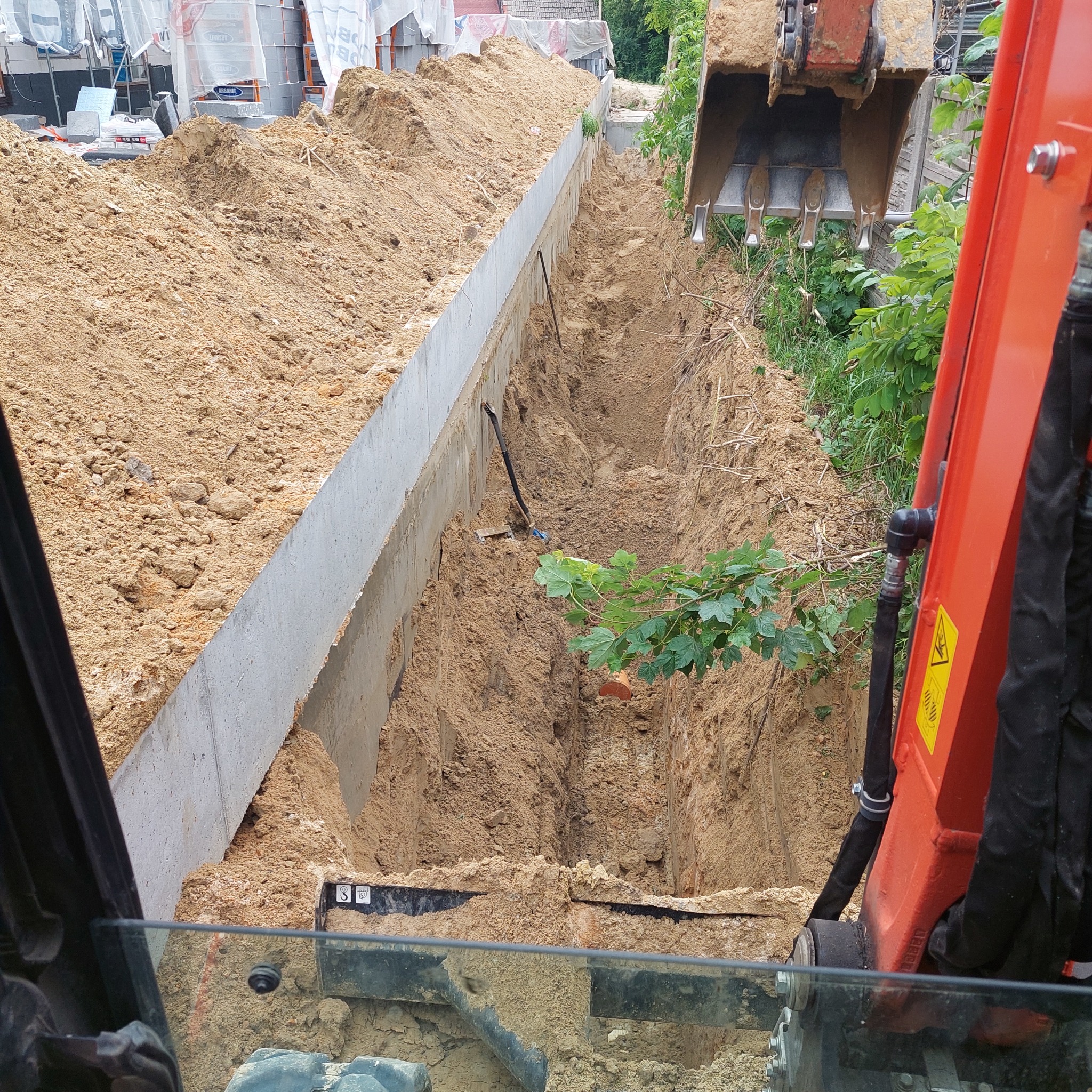 Wąski wykop z betonową ścianą, widok z kabiny koparki. Na dnie wykopu widoczne rury i gałąź. Koparka w trakcie pracy, ziemia usypana po bokach wykopu.