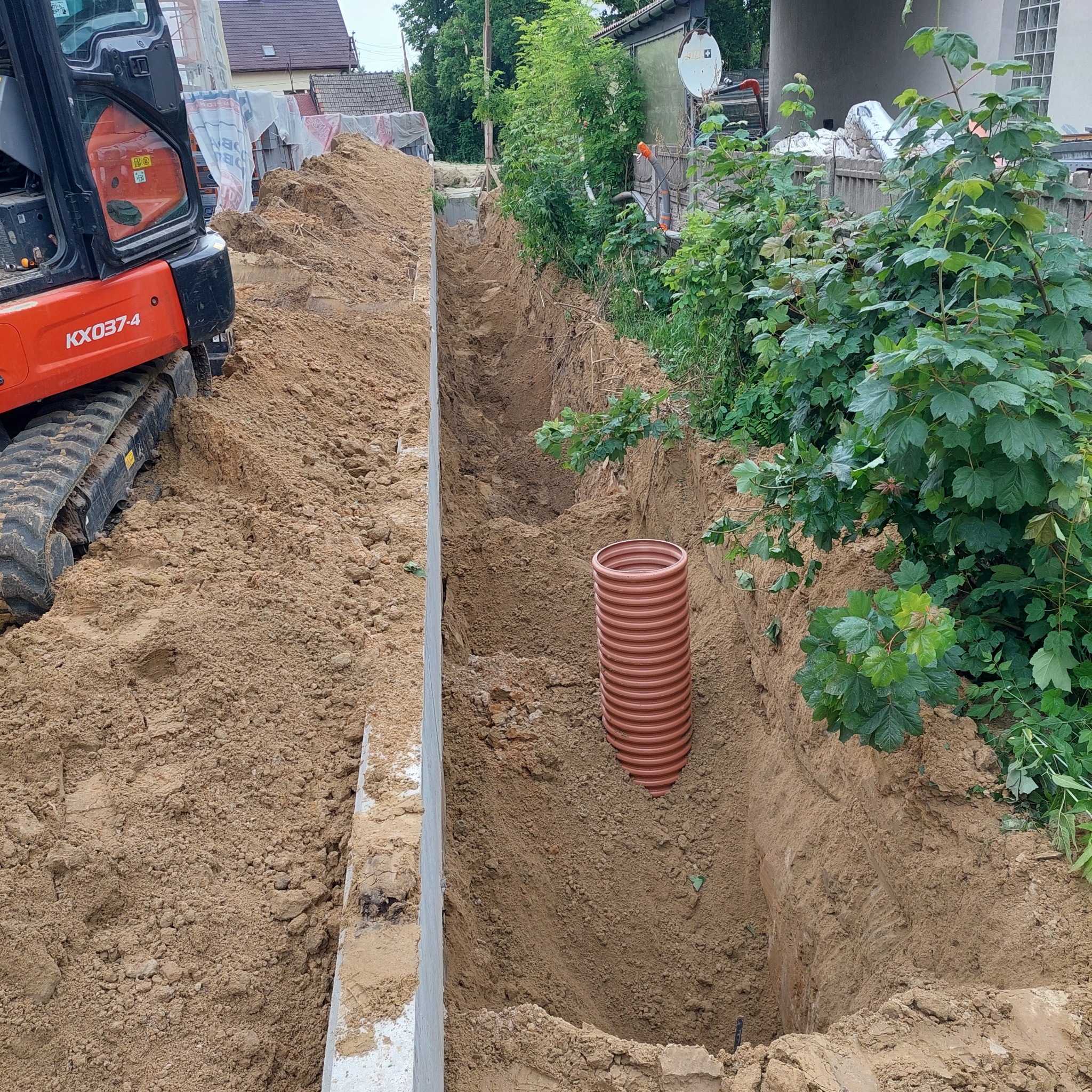 Wykopany wąski rów z pomarańczową rurą w środku, obok stoi mała koparka. Widoczne ślady pracy ziemnej i przygotowania do instalacji.