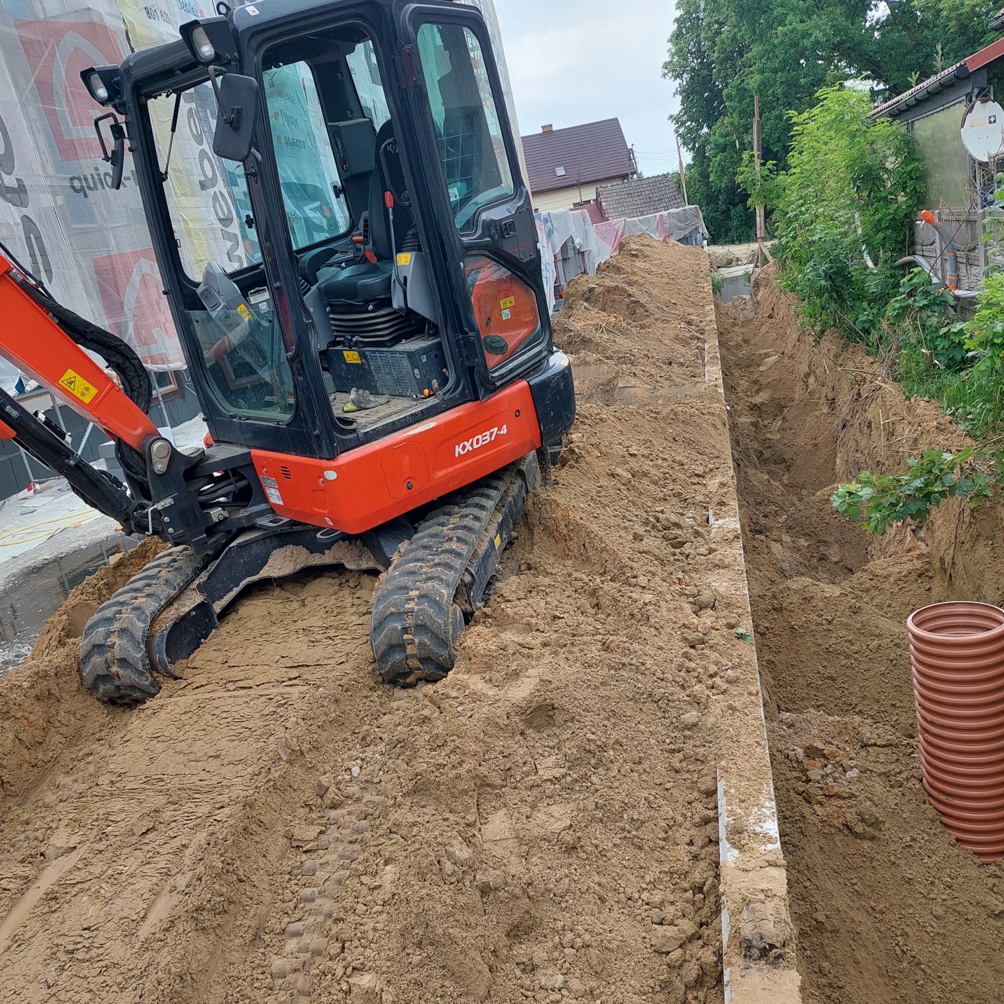 Pomarańczowa mini koparka Kubota KX037-4 na gruncie obok wykopu pod instalacje. W tle budynek mieszkalny i zieleń. Widoczna rura kanalizacyjna.
