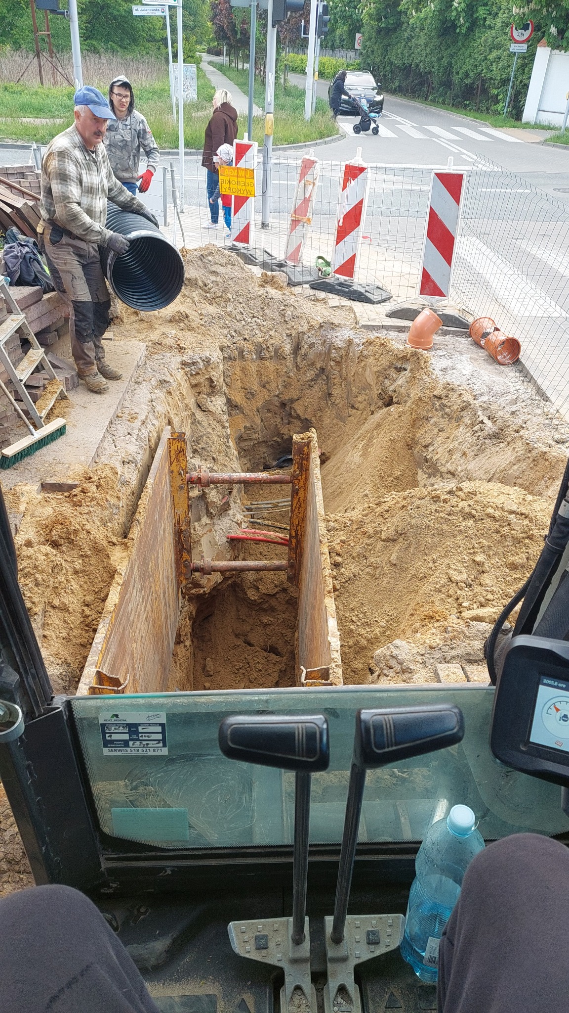 Głęboki wykop zabezpieczony szalunkiem stalowym. Robotnik w kasku i rękawicach umieszcza rurę w wykopie. Widok z kabiny koparki, w tle ulica z przejściem dla pieszych.