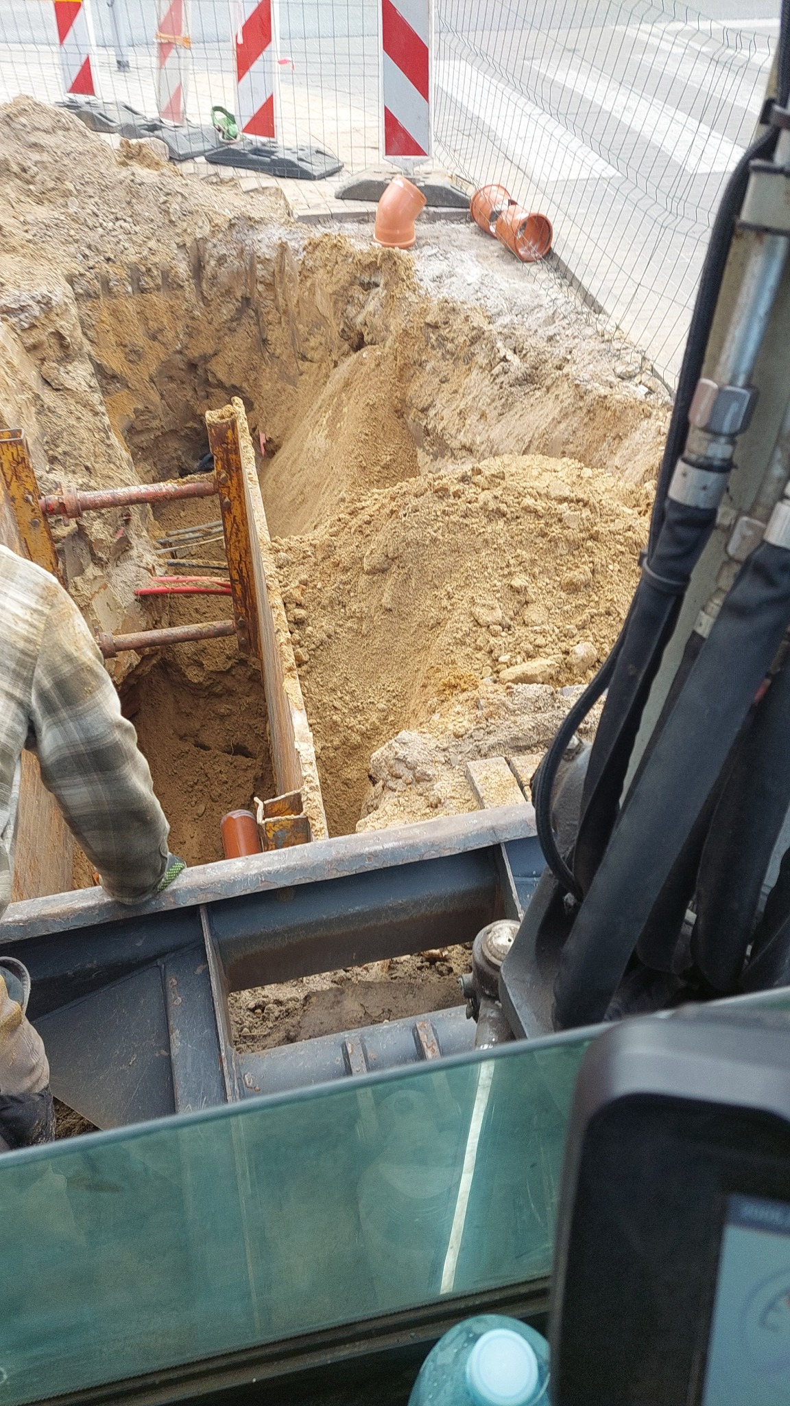 Głęboki wykop zabezpieczony szalunkiem stalowym, widoczne rury kanalizacyjne, fragment koparki i pracownik w koszuli w kratę. Prace ziemne w toku.