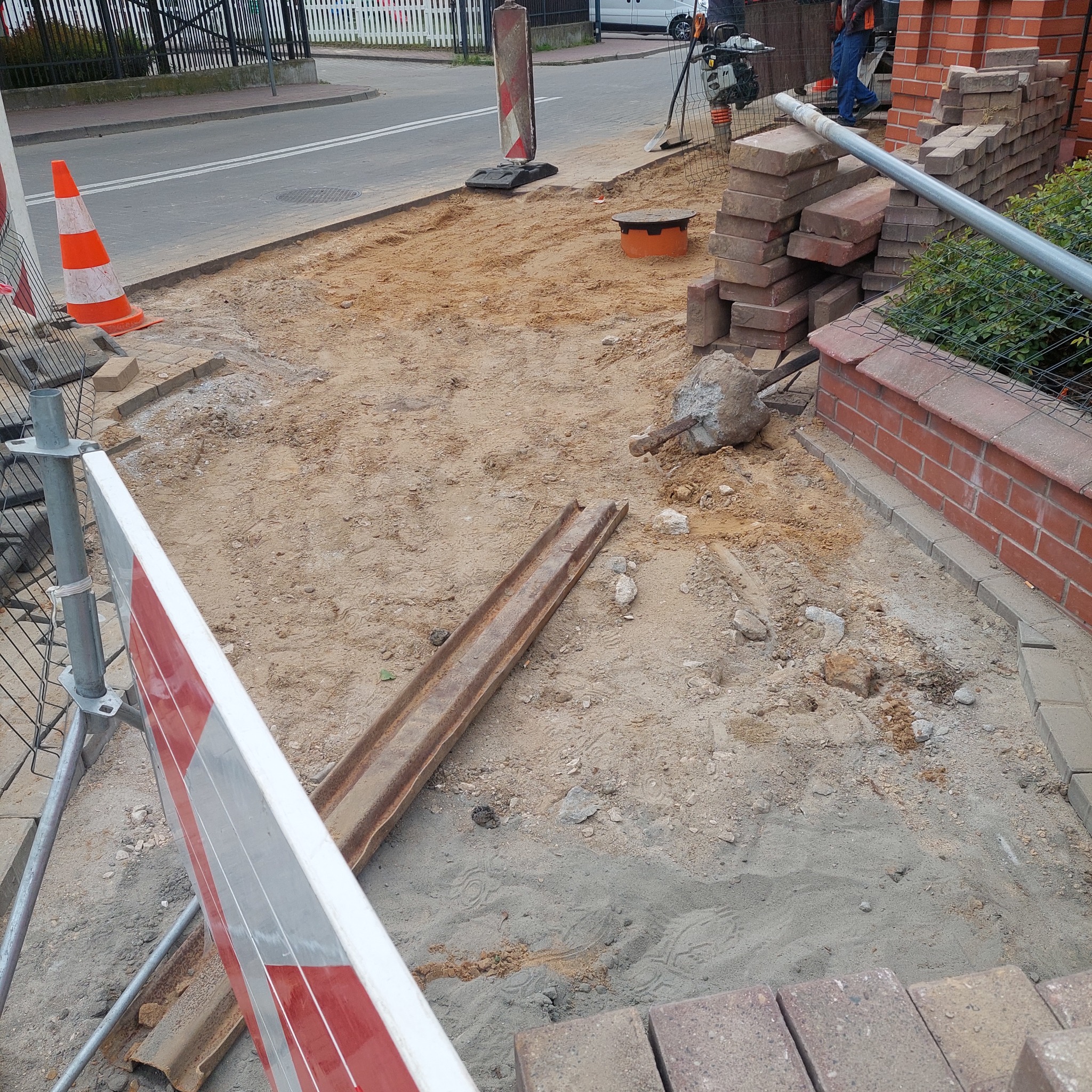 Przygotowanie podłoża pod kostkę brukową z widocznymi elementami zabezpieczającymi teren budowy, takimi jak bariery i pachołki. Częściowo ułożona kostka.