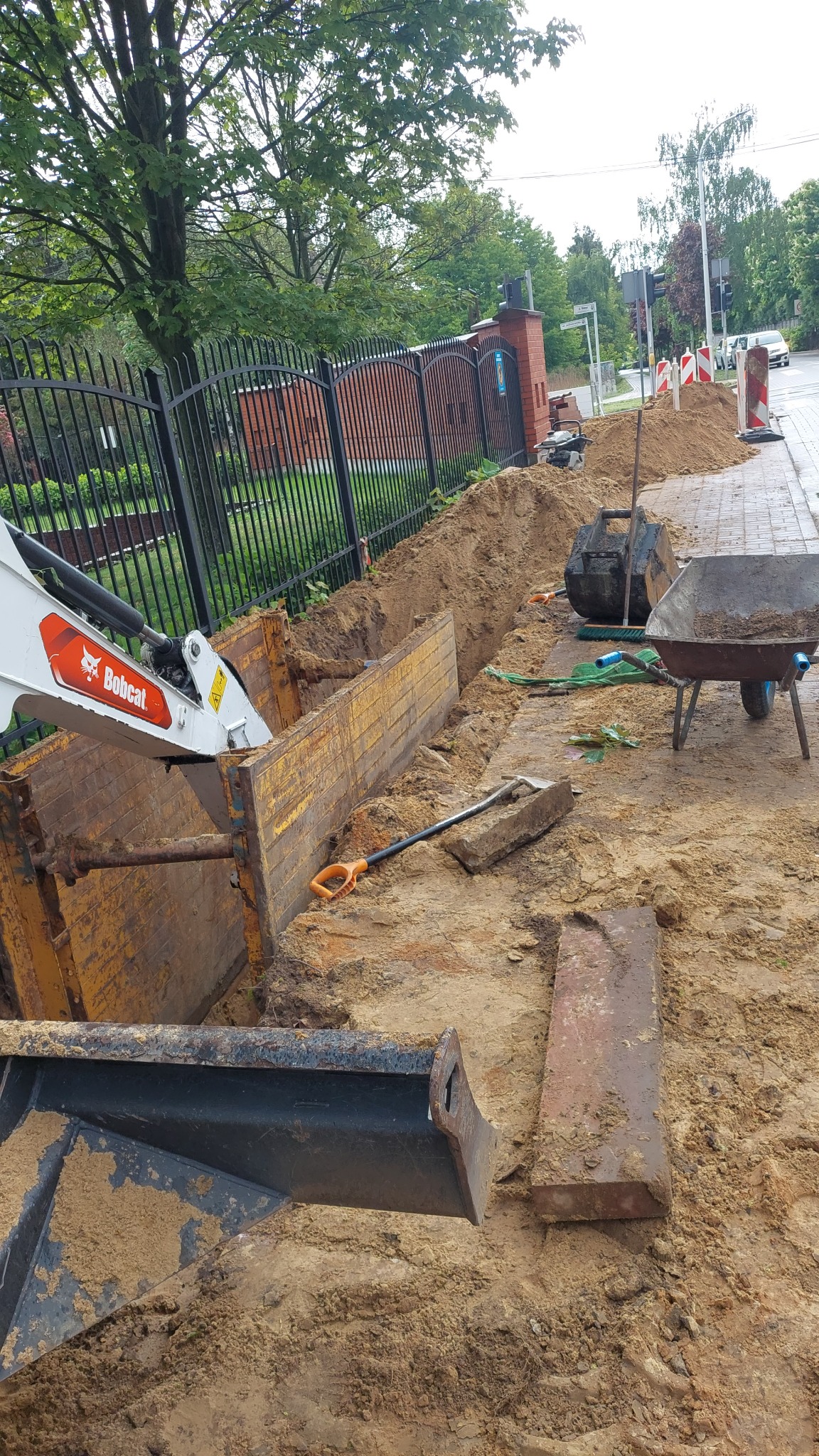 Wykop zabezpieczony szalunkiem, z widocznym ramieniem koparki Bobcat, łopatą i taczką. Prace ziemne wzdłuż chodnika i ogrodzenia z ceglanym słupkiem.