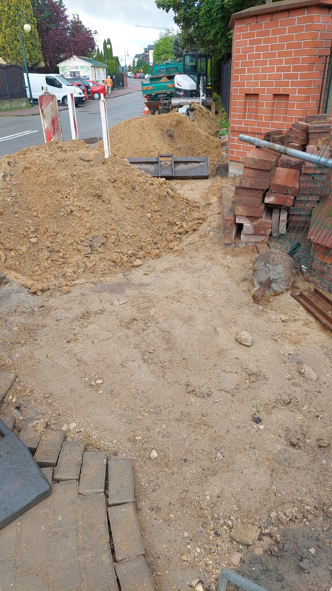 Przygotowanie terenu pod kostkę brukową w Regułach. Widoczny mini-ładowarka, sterta piasku i częściowo ułożona kostka. Prace ziemne w toku.